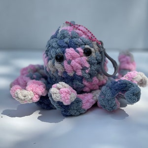 Puede incluir: Un juguete de peluche de pulpo de crochet hecho con hilo rosa, gris y blanco. El pulpo tiene grandes ojos negros y un clip de pl&aacute;stico gris sujeto a su cabeza.