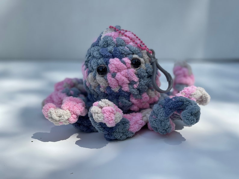 Puede incluir: Un juguete de peluche de pulpo de crochet hecho con hilo rosa, gris y blanco. El pulpo tiene grandes ojos negros y un clip de pl&aacute;stico gris sujeto a su cabeza.