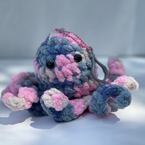 May include: A crocheted octopus keyring with pink, blue and white yarn. The octopus has large black eyes and a grey metal clasp.