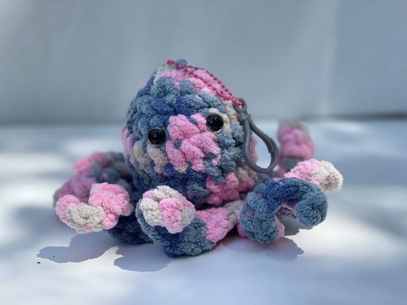 Puede incluir: Un llavero de pulpo tejido a crochet con hilo rosa, azul y blanco. El pulpo tiene grandes ojos negros y un cierre de metal gris.