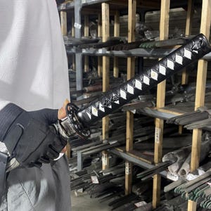 May include: A black katana sword with a white diamond pattern on the handle. The sword is being held by a person wearing black gloves. The sword is in front of a shelf with many other swords.