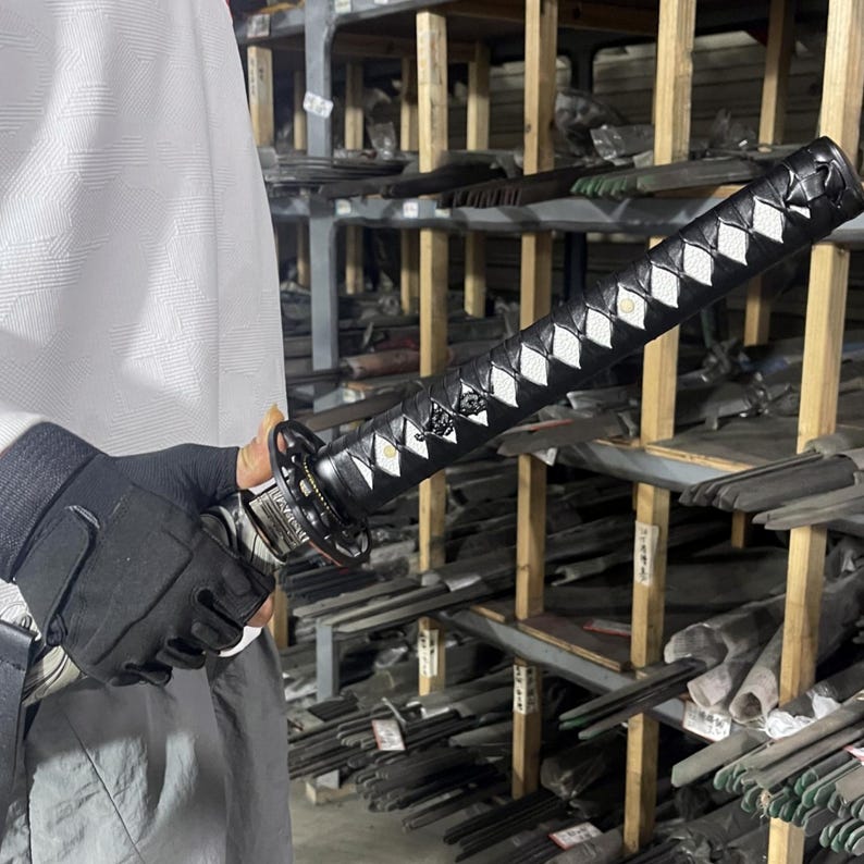 May include: A black katana sword with a white diamond pattern on the handle. The sword is being held by a person wearing black gloves. The sword is in front of a shelf with many other swords.