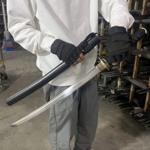 May include: A person wearing black gloves holds a silver katana sword with a black scabbard. The sword has a gold-coloured hilt and a decorative pattern on the blade.