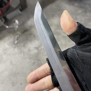 May include: A close-up of a silver katana blade with a slight curve. The blade is sharp and has a polished finish. The blade is held in a gloved hand.