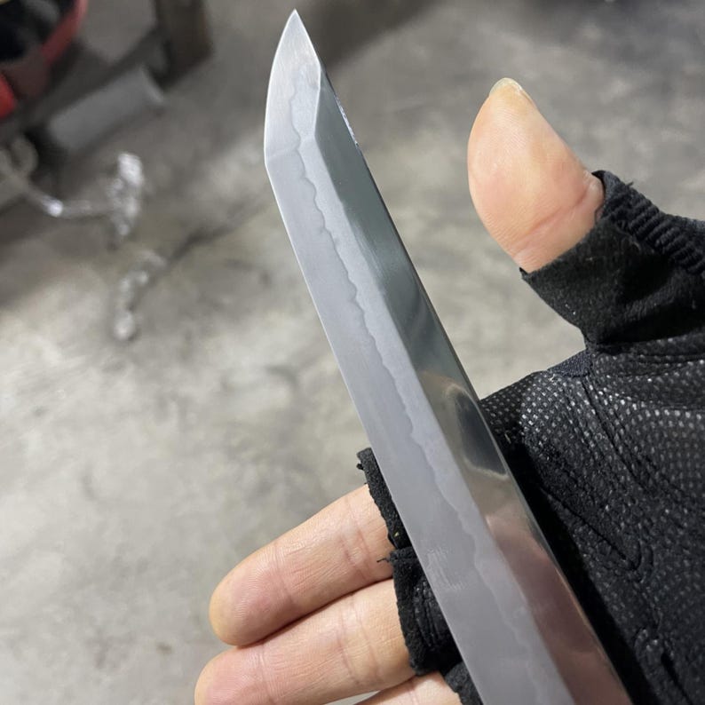 May include: A close-up of a silver katana blade with a slight curve. The blade is sharp and has a polished finish. The blade is held in a gloved hand.