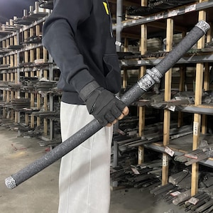 以下が含まれることがあります： 銀色の柄と複雑なデザインが施された黒い刀。刀は黒い手袋と黒いパーカーを着た人が持っている。刀は、他の刀でいっぱいになった棚の壁の前にある。