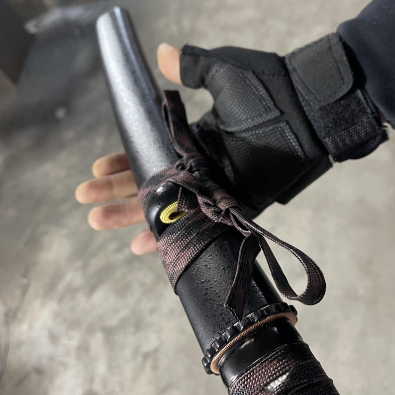 May include: A black sword scabbard with a brown and black braided wrap. The scabbard has a yellow metal ring and a black strap with a gold buckle.