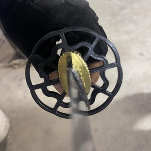 May include: Close-up of a black sword with a gold-coloured tsuba, a circular guard with a decorative design. The sword is held by a hand wearing a black glove.