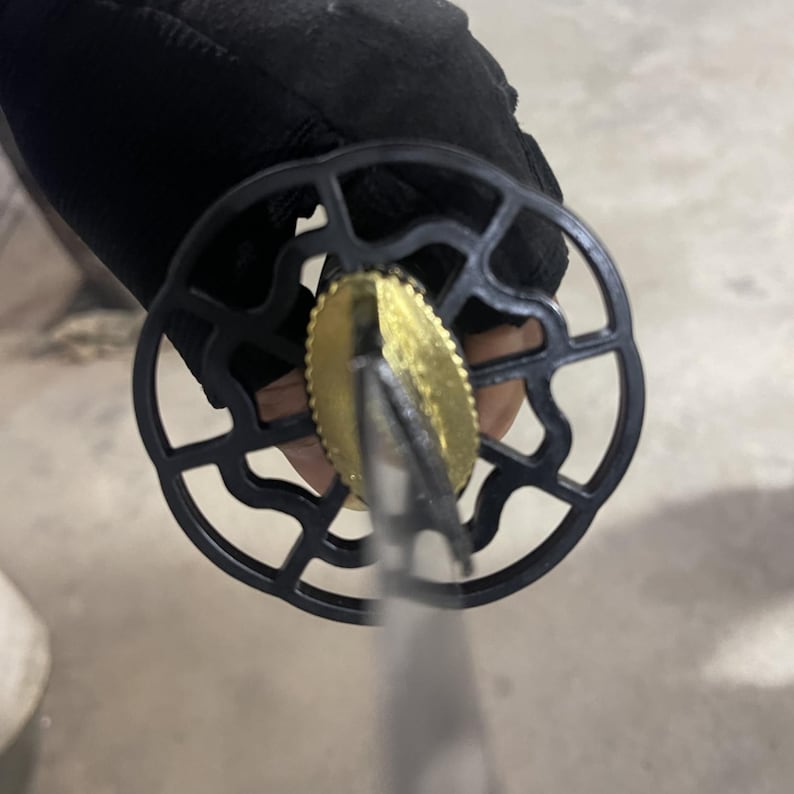 May include: Close-up of a black sword with a gold-coloured tsuba, a circular guard with a decorative design. The sword is held by a hand wearing a black glove.