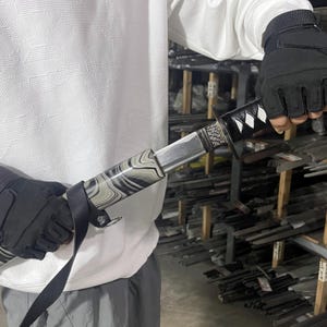 May include: A black and white patterned katana sword with a silver blade and a black handle. The sword is being held by a person wearing a white shirt and black gloves.