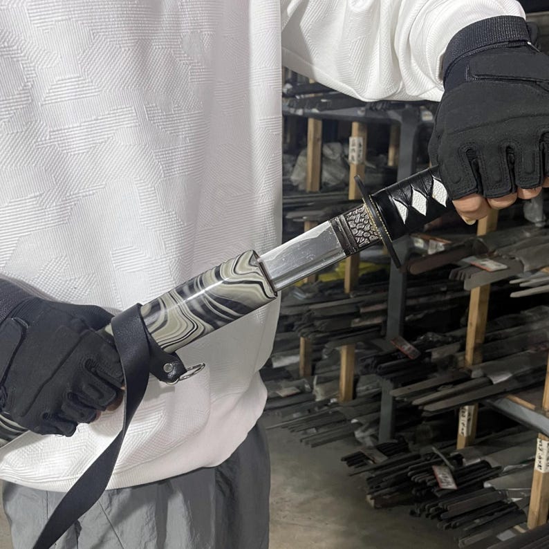 May include: A black and white patterned katana sword with a silver blade and a black handle. The sword is being held by a person wearing a white shirt and black gloves.