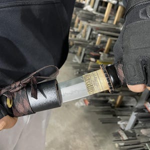May include: A close-up of a black and brown katana sword with a gold and black patterned handle. The sword is being held by a person wearing black gloves.