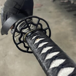 May include: Close-up of a black katana sword with a white and black braided handle and a black metal tsuba, or hand guard, with a decorative design.