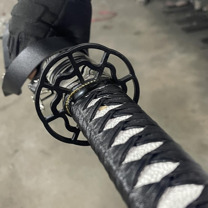 May include: Close-up of a black katana sword with a white and black braided handle and a black metal tsuba, or hand guard, with a decorative design.