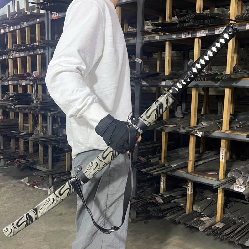 May include: A black and white patterned katana sword with a black leather handle and a black strap. The sword is being held by a person wearing a white sweatshirt and grey trousers.