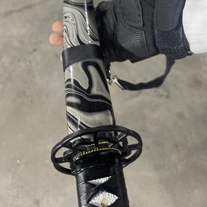 May include: A black and grey katana sword with a decorative handle and a black scabbard. The scabbard has a marbled pattern.