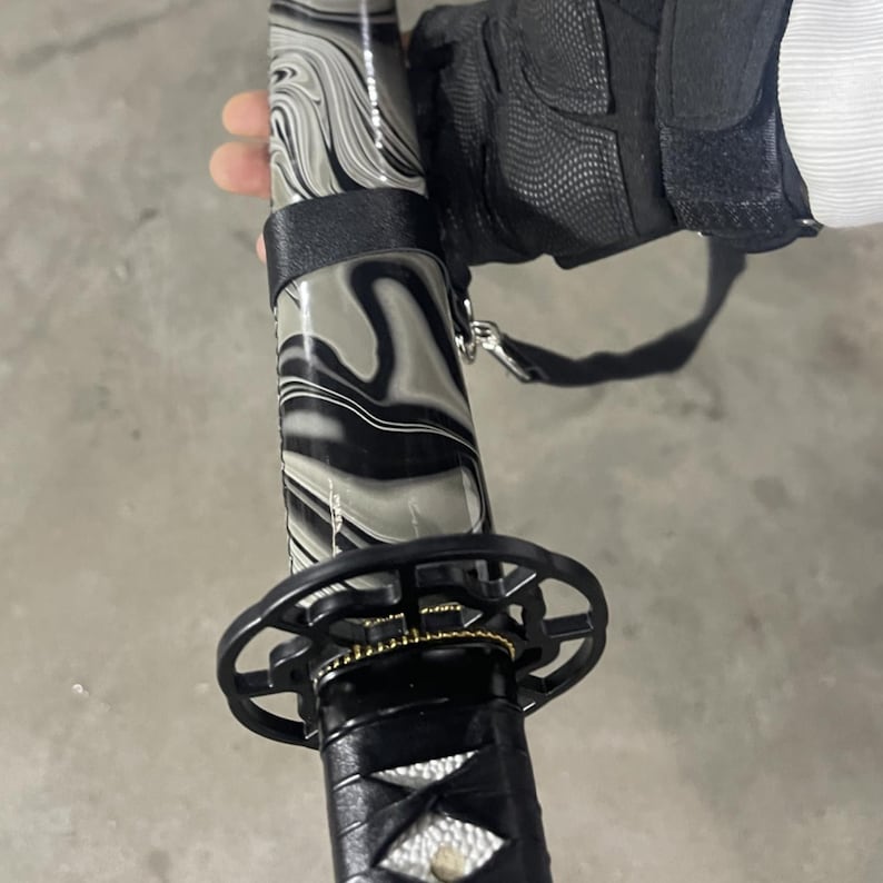 May include: A black and grey katana sword with a decorative handle and a black scabbard. The scabbard has a marbled pattern.