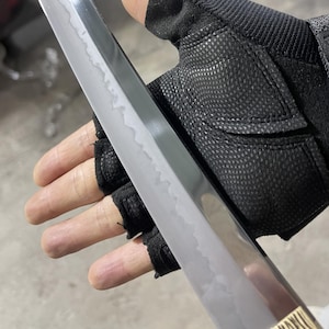 May include: A close-up of a silver sword blade with a textured surface. The blade is being held by a gloved hand.