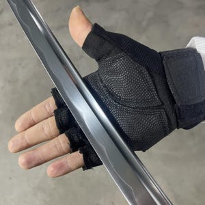 May include: A close-up of a silver sword blade with a black fingerless glove holding the blade. The blade has a slight curve and a textured surface.