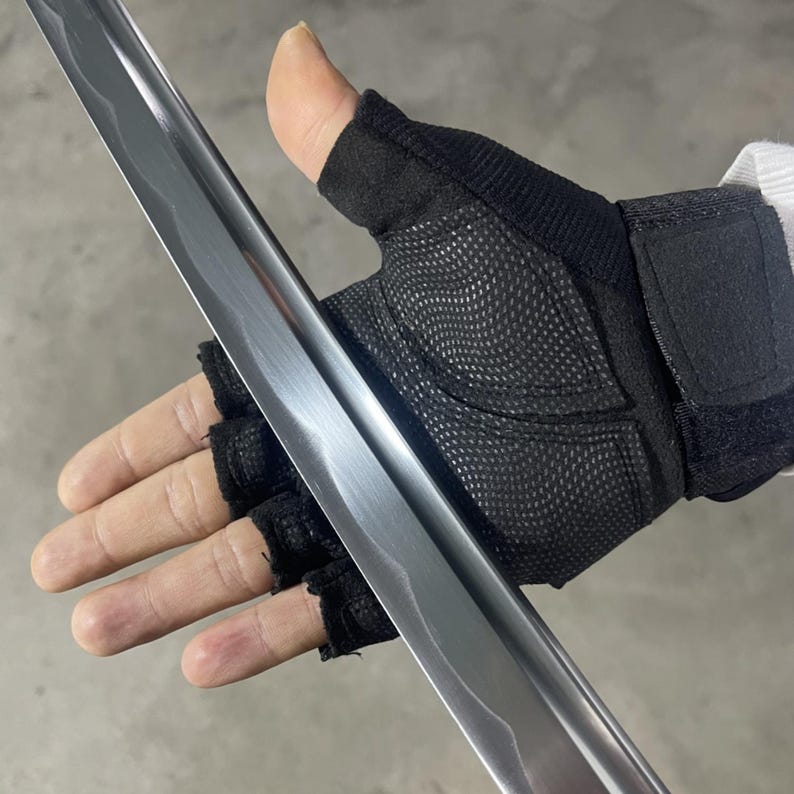 May include: A close-up of a silver sword blade with a black fingerless glove holding the blade. The blade has a slight curve and a textured surface.