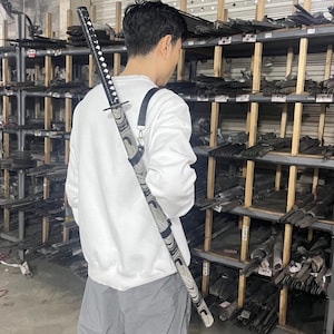 Pode incluir: Uma pessoa vestindo uma camisa de moletom branca está carregando uma espada katana preta e branca em uma bainha preta com uma alça sobre o ombro. A espada está em um depósito com prateleiras cheias de barras de metal.