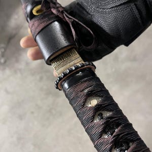 May include: Close-up of a black and brown sword handle with a white and black braided wrap. The handle is wrapped with a black leather sheath and has a decorative metal band.