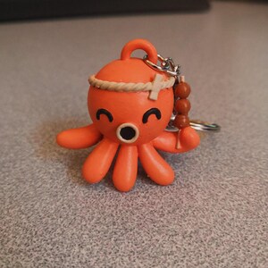 Peut inclure: Porte-clés en forme de pieuvre orange avec un bandeau en corde marron et des perles marron. La pieuvre a un visage souriant et tient une perle marron dans sa tentacule droite.