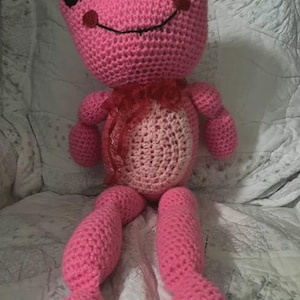 Könnte beinhalten: Eine handgefertigte rosa Häkelfroschpuppe mit schwarzen Augen und einem lächelnden Mund. Der Frosch hat einen weißen Bauch und ein rotes Band um den Hals. Die Beine des Frosches sind ausgestreckt und er sitzt auf einer weiß gemusterten Oberfläche.