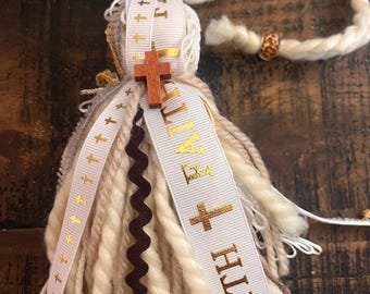 May include: A handmade tassel made from cream-coloured yarn, with a wooden cross and ribbons. The ribbons display the words "FAITH" and "ETERNITY" in gold lettering, along with small gold crosses. A decorative piece with a spiritual theme.