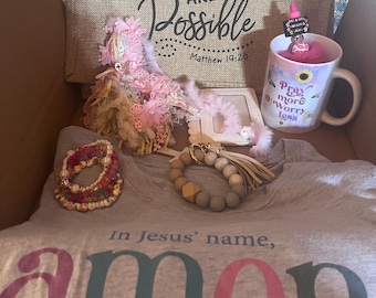 Peut inclure: Un t-shirt gris avec les mots "In Jesus' name, amen" imprimés en rose, vert et bleu. Le t-shirt est dans une boîte avec d'autres articles, dont un sac en toile de jute avec les mots "With God all things are possible Matthew 19:26", une tasse rose et blanche avec les mots "Pray more worry less", un bracelet en perles rose et blanc et une fleur rose et blanche.