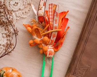 May include: A large green paperclip adorned with a decorative cluster of autumn-themed ribbons and fabric strips. The embellishments feature orange, red, and cream colours with patterns of pumpkins and leaves, accented by small orange pom-poms and yarn.