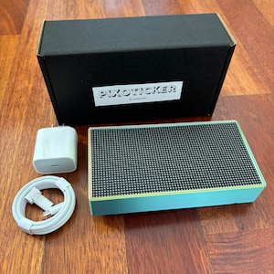May include: A mint green and black speaker with a black box, charger, and white cable. The speaker has a grid pattern on the front. The box is black with the text "PIXOTICKER" in white. The charger is white.