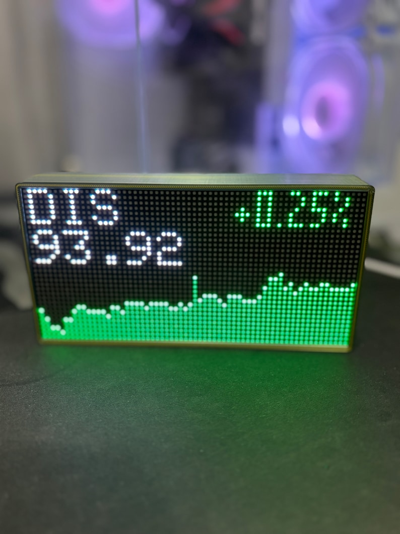 May include: A rectangular LED display shows stock market data. The display shows "DIS" and "93.92" in white, with a green graph and "+0.25%" on the right. The device has a dark frame.