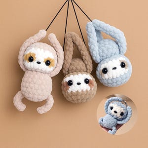 Op de afbeelding: Drie gehaakte luiaardornamenten in lichtroze, bruin en blauw. Elke luiaard heeft een wit gezicht, zwarte ogen en een kleine, bruine neus. De luiaards hangen aan zwarte touwtjes, met een klein luiaardornament rechtsonder.