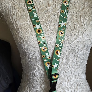 May include: Green lanyard featuring sunflowers, white stars, and the text "I am Autistic." It has a black plastic clasp and a metal clip for attaching keys or an ID badge. The lanyard is approximately 80 cm long.