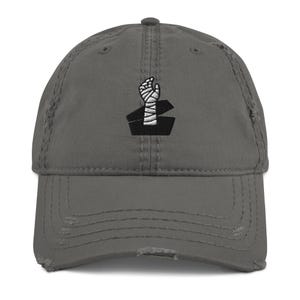 May include: Distressed gray baseball cap featuring an embroidered design of a wrapped fist emerging from a coffin. The cap has a pre-curved visor and a distressed finish, giving it a vintage look.
