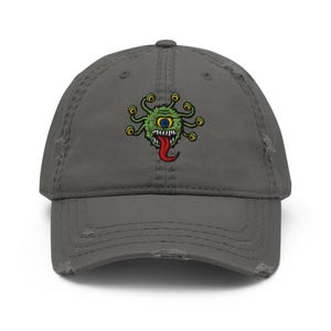 Pode incluir: Boné de beisebol cinza desgastado com um monstro verde bordado. O monstro tem um olho grande, vários olhos menores e uma língua vermelha. O boné tem um visual envelhecido com bordas desgastadas.
