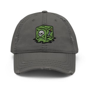 Könnte beinhalten: Verwaschene graue Baseballkappe mit einem gestickten Design eines grünen Würfels mit Totenkopf, Messer und Tropfeffekt. Die Kappe hat einen Used-Look mit ausgefransten Kanten.