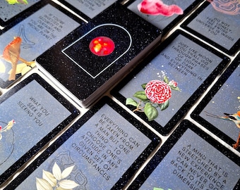 May include: A tarot card deck with a black glittery border and a red orb design. Each card features illustrations of flowers, birds, and butterflies, with inspirational quotes in white text. The cards are arranged on a grey surface.