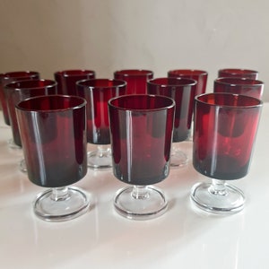 Pode incluir: Conjunto de 12 copos de shot de vidro vermelho rubi com hastes e bases de vidro transparente. Os copos estão dispostos em forma de escada sobre uma superfície branca.