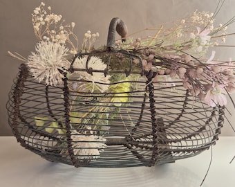 Puede incluir: Una cesta decorativa de alambre con asa, llena de flores y follaje secos. La cesta es de forma ovalada y está hecha de metal oscuro. El arreglo incluye flores secas blancas, rosas y amarillas.