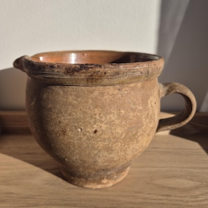 May include: A rustic, antique-style pottery jug with a handle and spout. The jug is a warm, earthy brown color with a slightly rough texture. The interior has a reddish-brown glaze. The jug sits on a wooden surface.