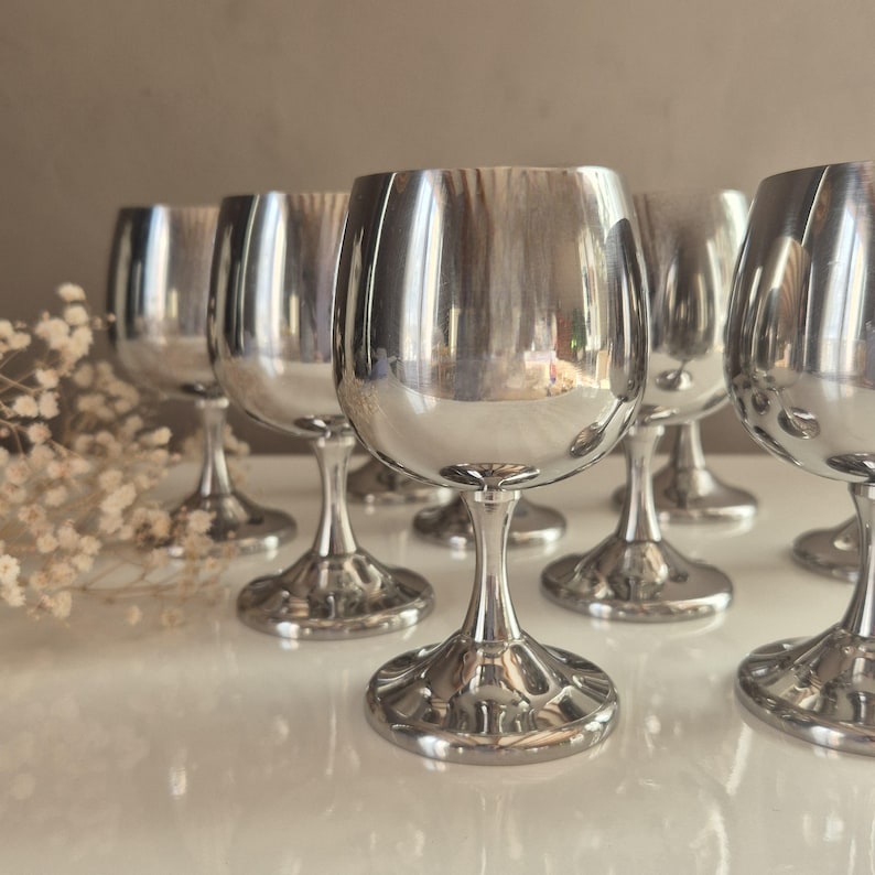 Puede incluir: Un juego de ocho copas de vino plateadas con una copa redonda y un tallo alto. Las copas est&aacute;n dispuestas en semic&iacute;rculo sobre una superficie blanca.