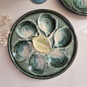 Puede incluir: Un plato de ostras de cerámica verde y blanco con seis hendiduras individuales para servir ostras. El plato tiene un borde elevado y un diseño decorativo de borde de cuerda.