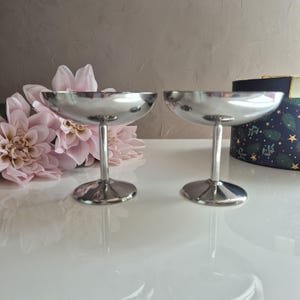 Peut inclure: Deux coupes à champagne argentées avec un large bol peu profond et une fine tige. Les verres sont présentés sur une surface blanche, avec des fleurs roses et une boîte décorative en arrière-plan. Les verres mesurent environ 10 cm de haut.
