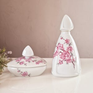 Può includere: Un set di due contenitori in porcellana bianca con motivi floreali rosa. Il contenitore più grande ha la forma di una bottiglia con un coperchio appuntito. Il contenitore più piccolo è una ciotola rotonda con un coperchio appuntito. Entrambi i contenitori hanno un motivo floreale con fiori rosa e foglie verdi.