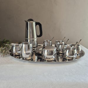 Puede incluir: Un juego de café de acero inoxidable sobre una bandeja ovalada. El juego incluye una cafetera, varias tazas pequeñas, un azucarero con tapa y cucharillas. El juego se exhibe sobre un mantel blanco.