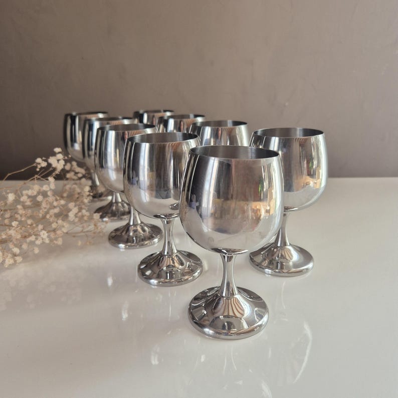 Puede incluir: Conjunto de diez copas de vino de metal plateado con una copa redonda y un tallo. Las copas est&aacute;n dispuestas en una fila sobre una superficie blanca.