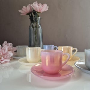 May include: A set of iridescent teacups and saucers in pastel colours, including pink, yellow, blue, and white. The cups are on a white surface, with a vase of pink flowers behind them. The teacups have a pearlescent finish.