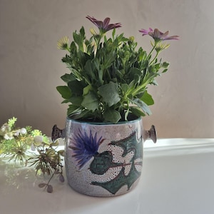 Peut inclure: Un pot en céramique avec un motif de chardon vert, blanc et bleu. Le pot a deux anses et est rempli d'une plante verte avec des fleurs violettes.